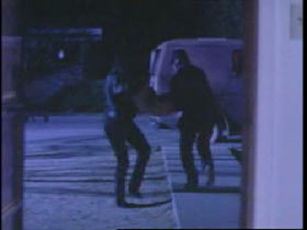 Two men dressed in black carrying a woman to a large white van.