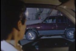 A man in the driver seat of a car staring out his window at a maroon SUV parked across the street.