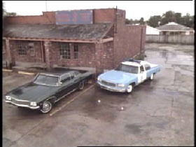 A black sedan parked infront of a brick building, a cop car is parking beside it.