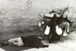 A homeless man sleeping on the floor against a brick wall. He's next to a shopping cart full of trash