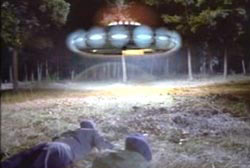 Two airmen laying on the ground, looking up at a hovering saucer with many lights