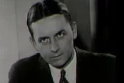 Eliot Ness sitting on a chair staring at the camera in a black suit.